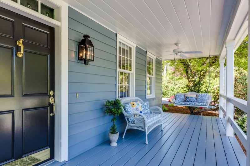 Covered front porch of a blue clapboard house, with a wicker chair and a hanging porch swing