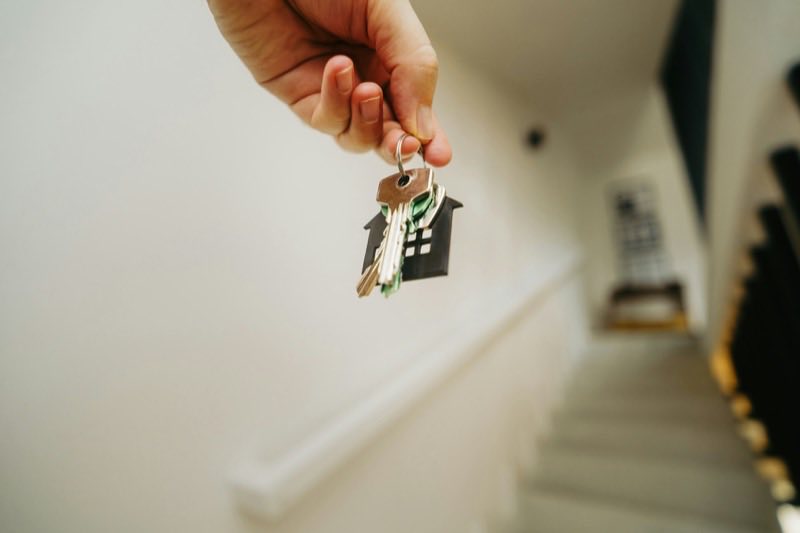 A hand holding house keys on a house-shaped keyring at the foot of a staircase