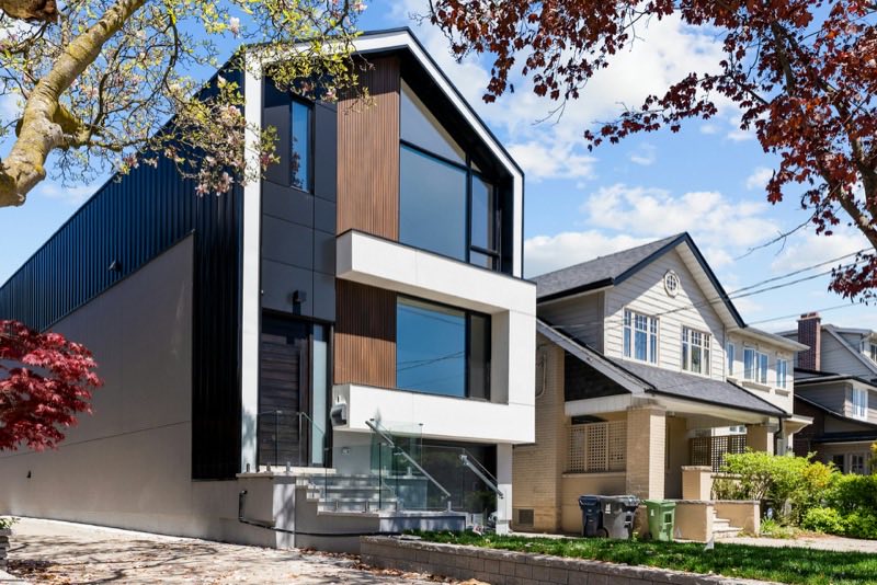 A modern infill house clad in wood and dark metal, standing between older neighboring homes