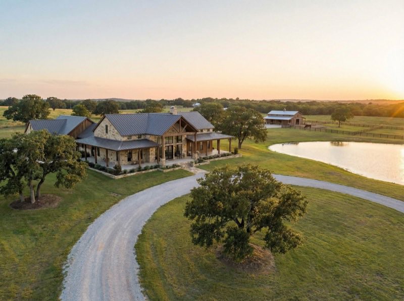 Aerial view of a large country house with a wraparound porch, a curving gravel drive and a pond