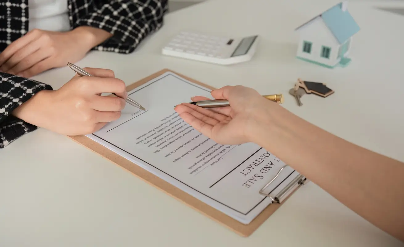 Two people at a desk reviewing a purchase and sale agreement on a clipboard, beside keys and a model house