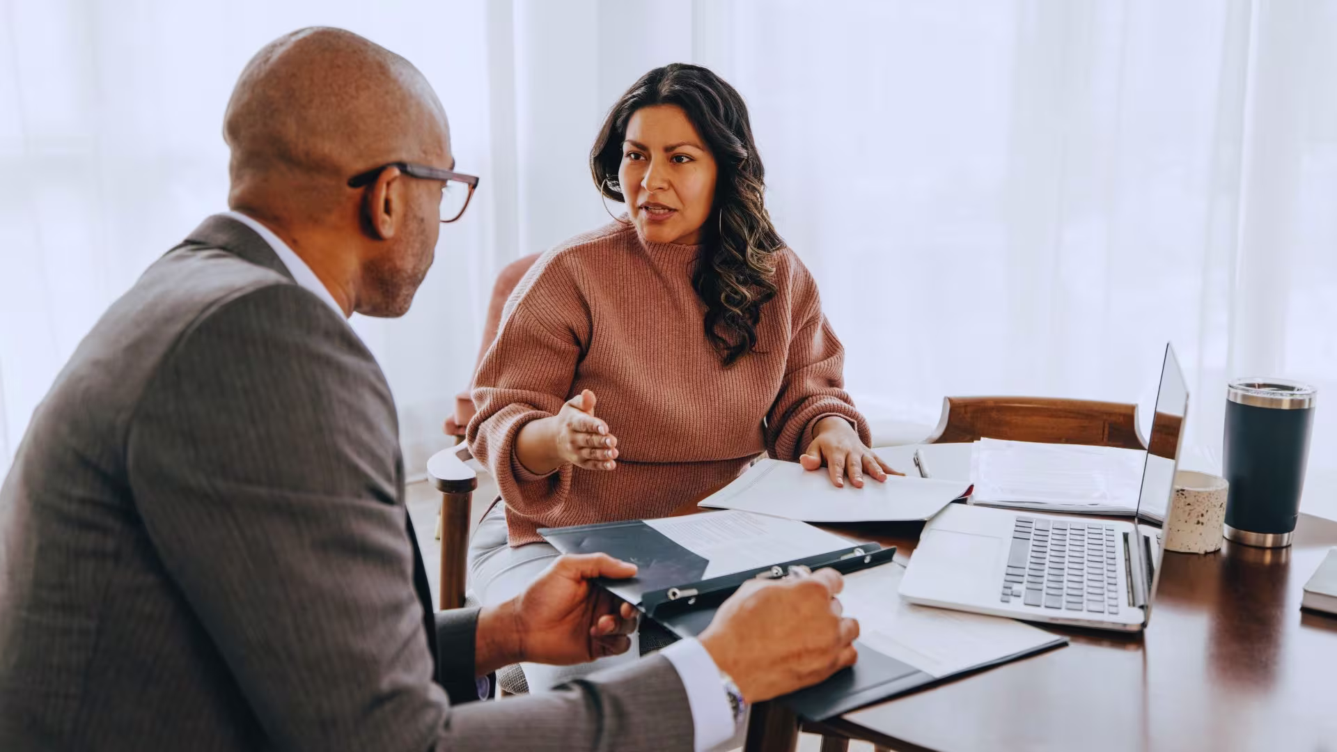 A loan officer and a client talking across a desk, with a laptop and paperwork between them