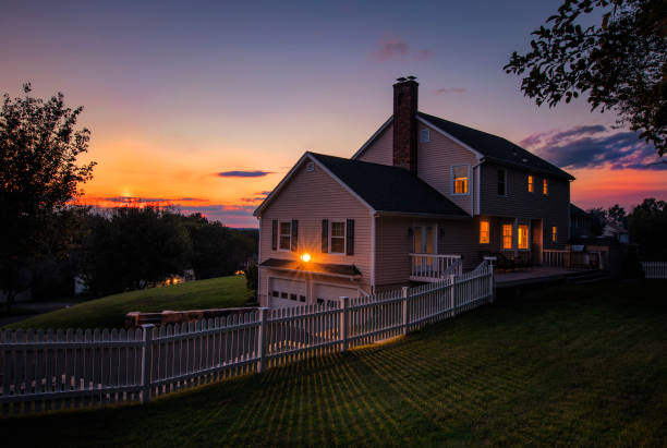 A two-story house behind a white picket fence, windows lit, with an orange sunset beyond it