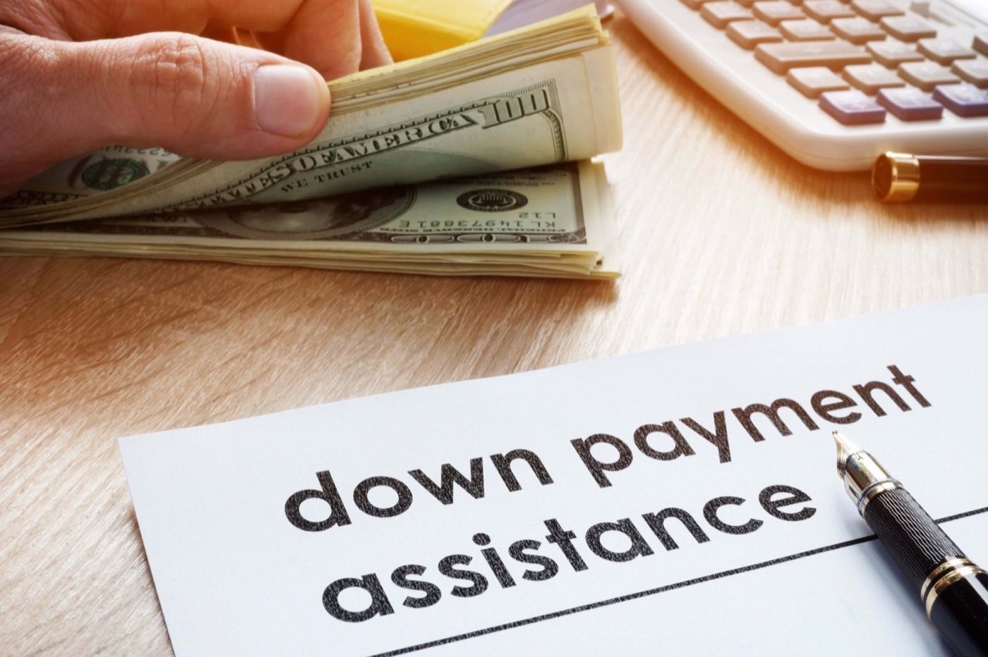 A hand holding a stack of cash beside a calculator and a document headed down payment assistance