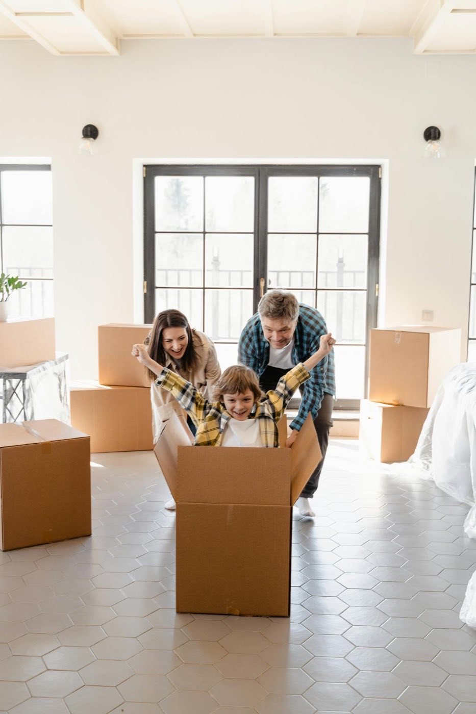 Two parents pushing a child in a cardboard box across an empty room on moving day, packing boxes stacked behind them