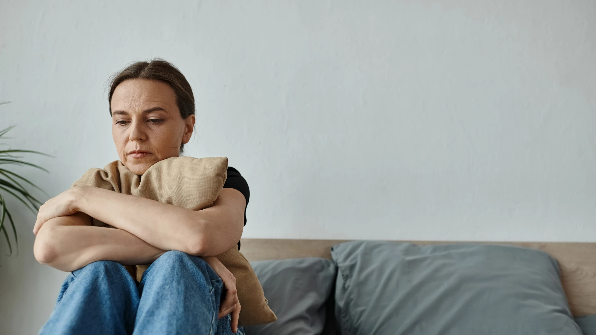 A person sitting on a bed hugging a cushion, looking away with a worried expression