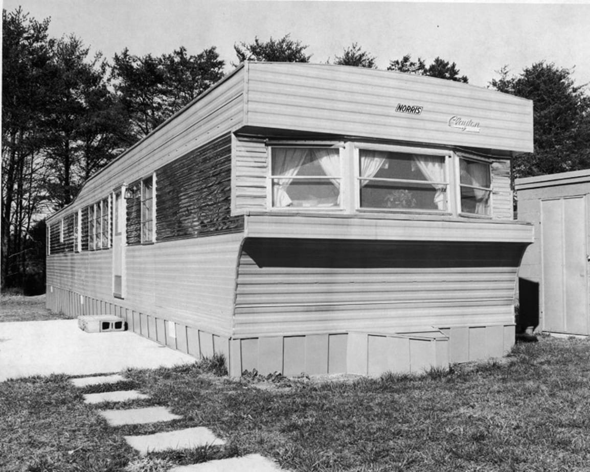 A black-and-white photograph of a 1960s single-wide mobile home with an awning over the front window, set on block piers