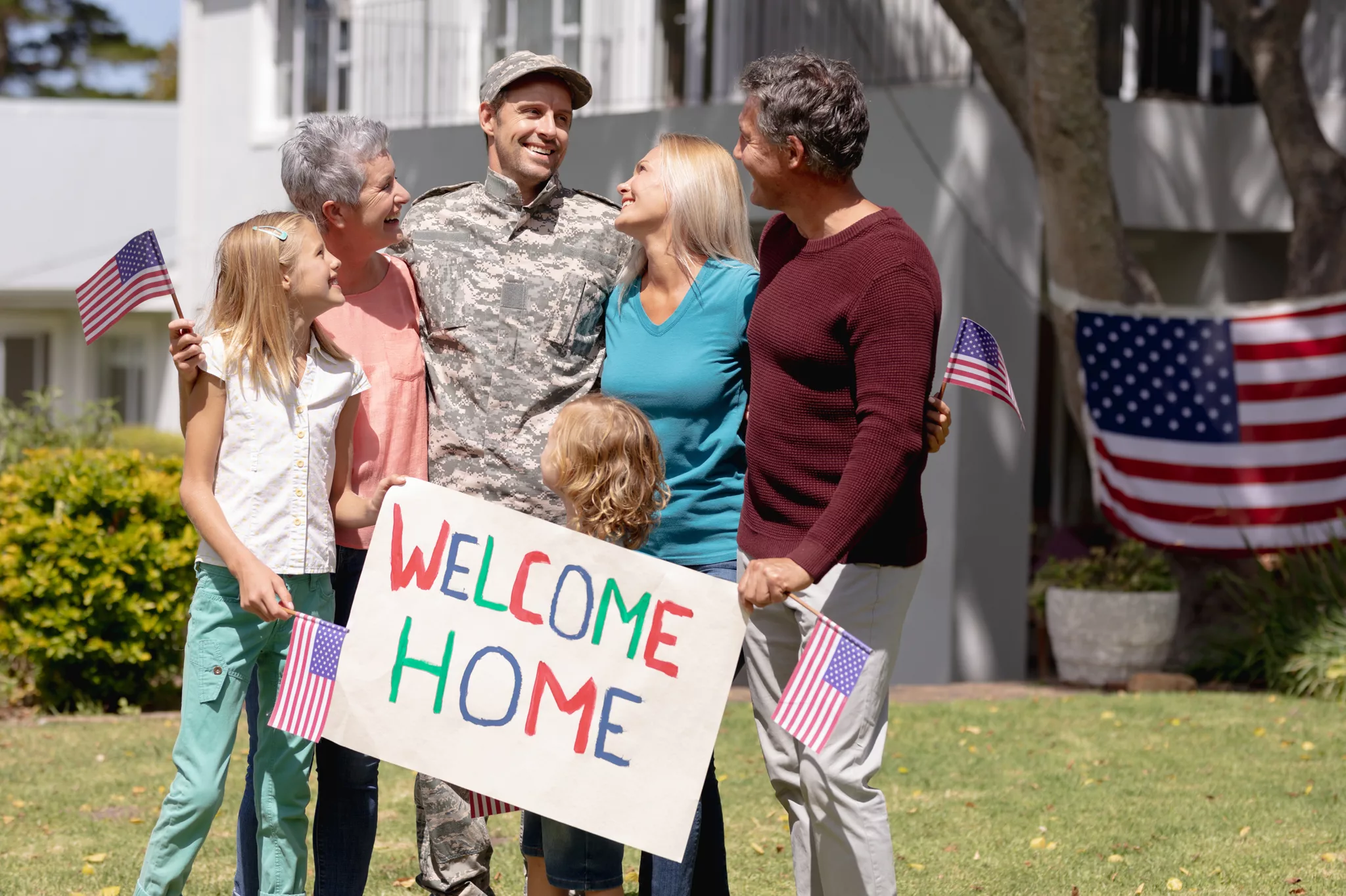 A family greeting a service member in camouflage uniform, holding a hand-lettered Welcome Home sign and small US flags